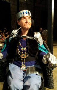 Doug is sitting in a wheelchair wearing a king's costume with a crown, necklace and a cape. Doug is looking upwards to a light.
