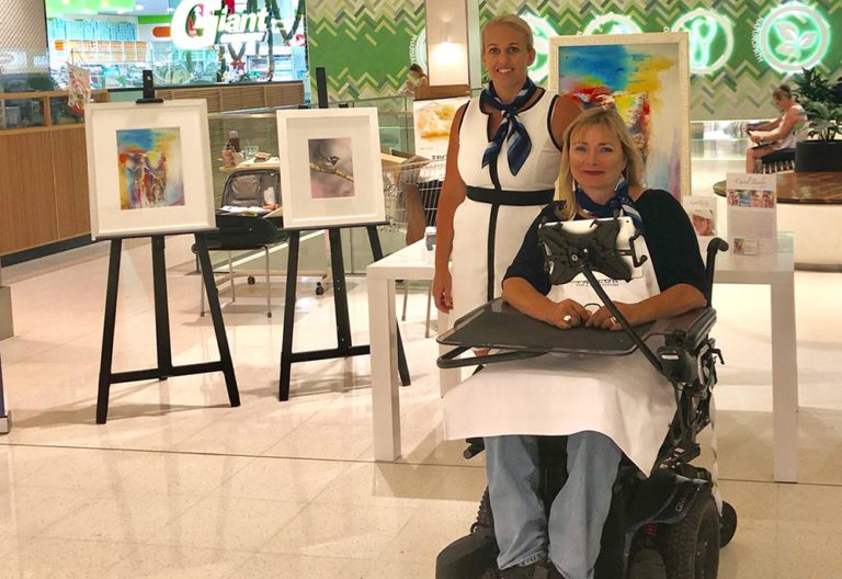 Two women, one in wheelchair, and one standing. Easels arranged behing showing watercolour artwork