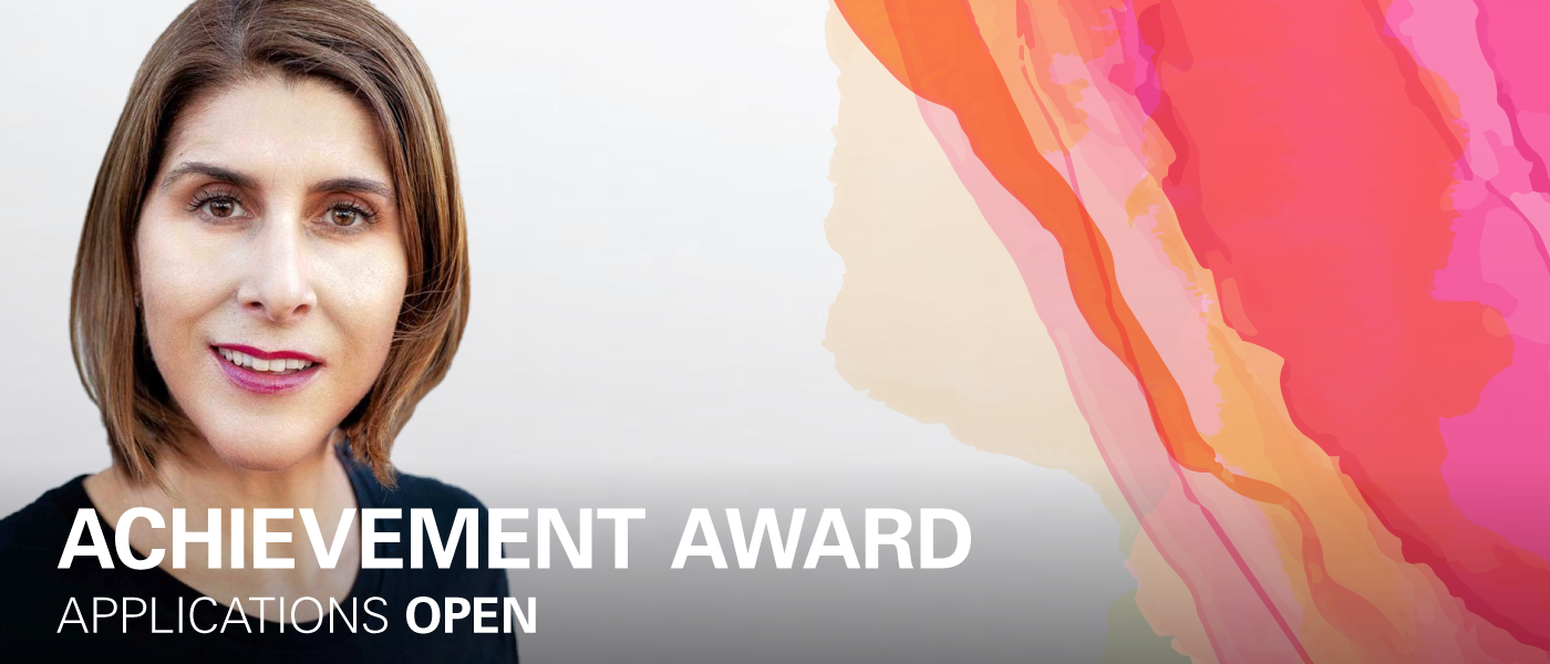 A woman with short brown hair is standing in front of a white background looking at the camera smiling. On the top right hand corner is a watercolour graphic in pinks and yellows. Text reads on the bottom of the image saying "Achievement Award - Applications Open"