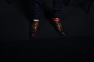 A pair of feet wearing brown shoes, red and white socks and navy pants on a black background