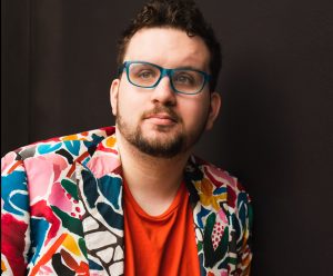 Oliver is standing in front of a black background wearing blue glasses, an orange shirt and a colourful blazer. He is looking slightly off from the camera lens with a calm expression.