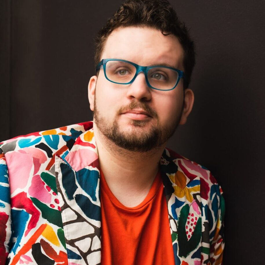 Oliver is standing in front of a black background wearing blue glasses, an orange shirt and a colourful blazer. He is looking slightly off from the camera lens with a calm expression.