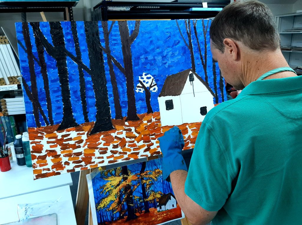 A person in a green shirt with short brown hair is painting. The painting is mostly complete and takes up most of the photograph. It depicts a white house in the woods with a dark blue sky and autumn-coloured leaves on the ground