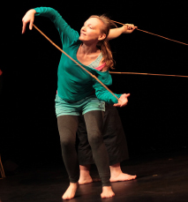 Performing artist Megan Louise West is wearing a long sleeved green shirt and black tights. The photo is taken of her mid-performance where she is dancing with a stick in between her hands.