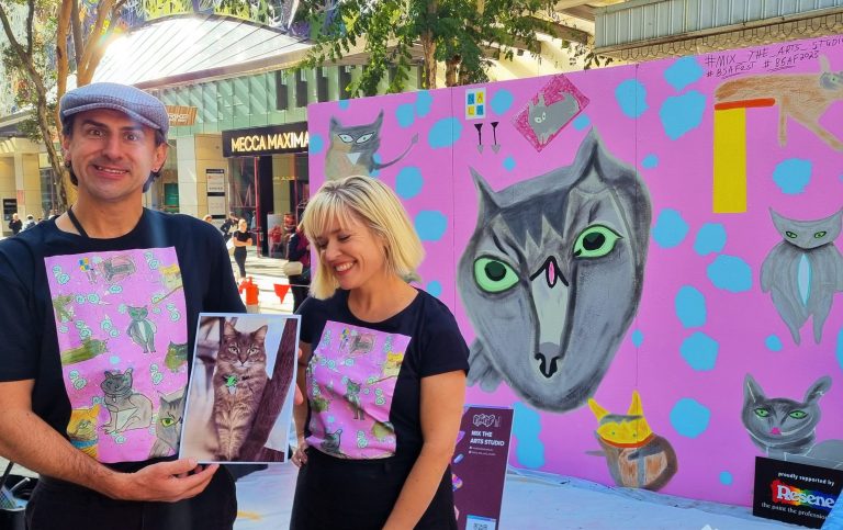 Tommy and Jasmin smiling at the camera holding a photo of a cat with a cat mural behind them