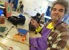 Paul is sitting at a table at one of the Access Arts visual art workshops. He has thick gray-ish hair and a purple smock on. In front of him is his work which appears to be multicoloured yarn weaved to depict an image.