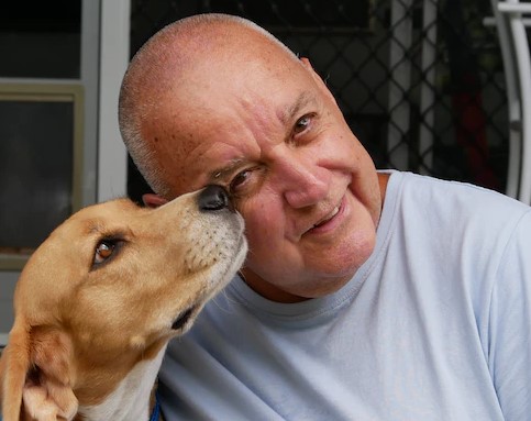 A man, George Parkyn wearing a light grey t-shirt sitting with his therapy dog 'Baxter' who is brown and white.
