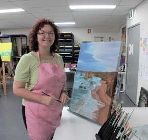 Kerry smiling for the camera in front of her artwork in an artist studio. She is wearing a green shirt with a pink apron over the top and her artwork is a beach setting.