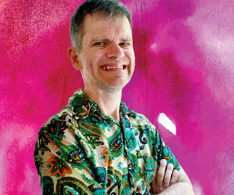 A man stands in a colourful t-shirt with his arms crossed. He smiles towards the camera. The background of his image is removed and he is placed on top of a vibrant pink and purple watercolour background.