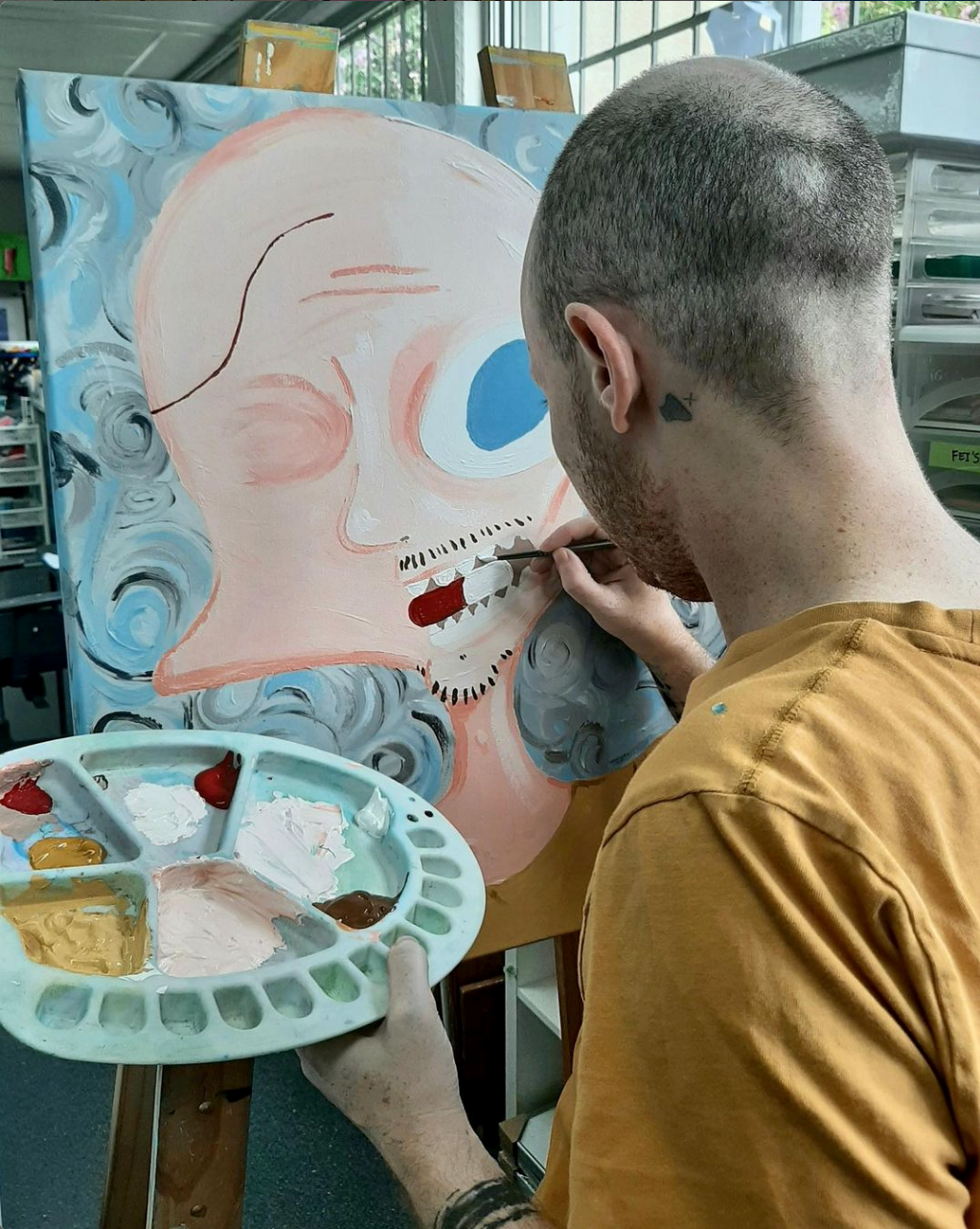 Jesse is painting on his easel. His back is to the camera, he is wearing a mustard coloured t-shirt and is holding a paint pallet in his hands. 