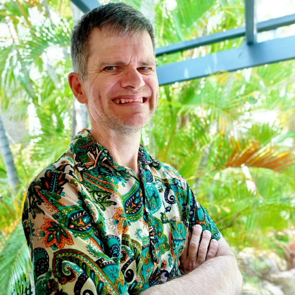 2024 Access Arts Achievement Award Winner - Michael Russell. Michael is facing the camera with his arms crossed. He is wearing a colourful Hawaiian shirt and he is smiling at the camera. He is standing outside amongst a lush garden.