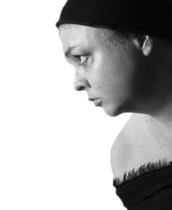 Black-and-white profile portrait of a woman with a shaved head, wearing a black beanie and an off-the-shoulder black top with a raw, fringed hem. She looks downward in three-quarter profile against a plain white background, in dramatic, high contrast lighting.