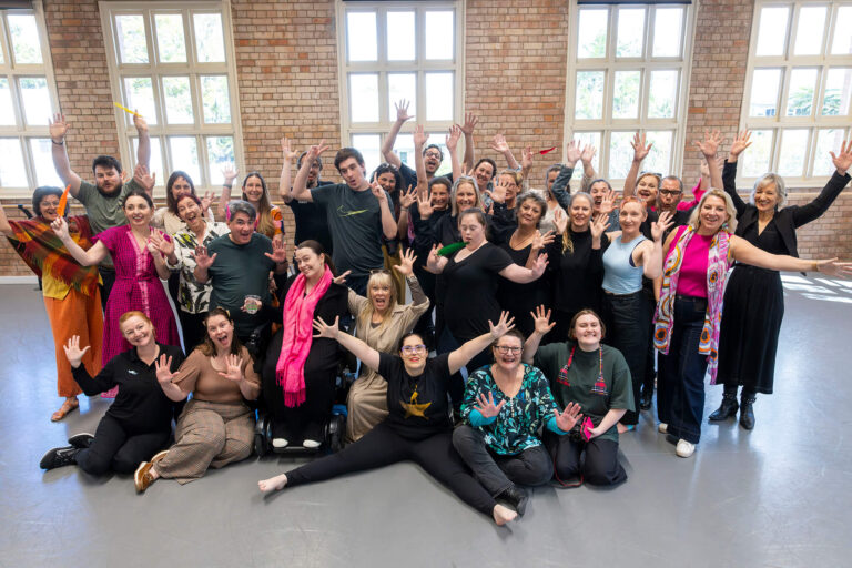 A group of artists standing together for a group photo. Some are standing, others are kneeling or sitting in a wheelchair. Everyone has their arms up with happy and excited expressions on their faces.