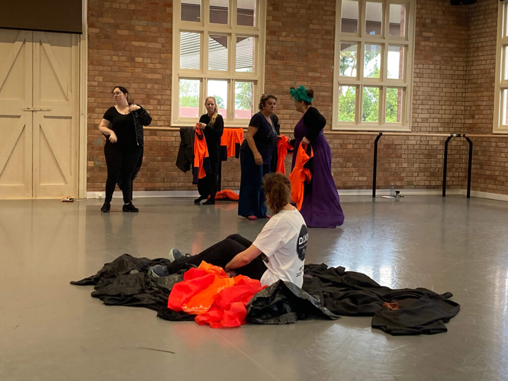 Performers on stage in a theatre space. One person looking away with black and orange pieces of material around them, while 4 others stand in the background interacting with each other.